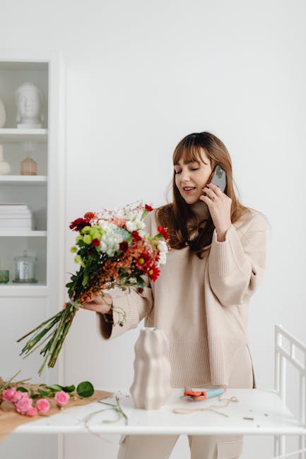 A young woman with long brown hair and bangs, dressed in a beige sweater, is holding a colorful bouquet of fresh flowers including red, pink, white, and green blooms with greenery accents. She is smil