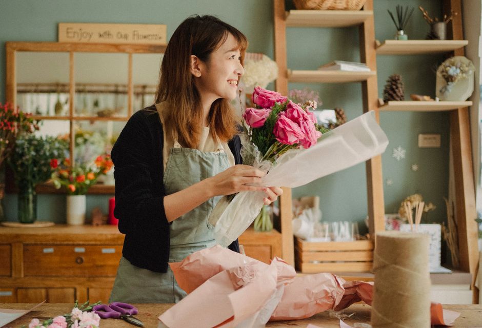A woman in a florist shop holding a bouquet of vibrant pink roses and soft pink lisianthus, wrapped in delicate white tissue paper, while smiling. She is wearing a beige apron over a dark top, and is