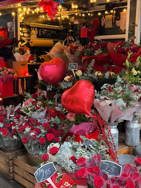 A display of vibrant floral arrangements featuring red roses, white gypsophila, and eucalyptus foliage, artistically arranged in baskets and wrapping paper. Heart-shaped red foil balloons are included