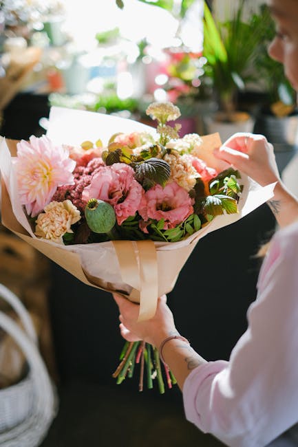 A person holding a large bouquet of fresh flowers wrapped in beige and white paper, featuring pink and white peonies, carnations, and greenery, with some small white blossoms. The bouquet is arranged 
