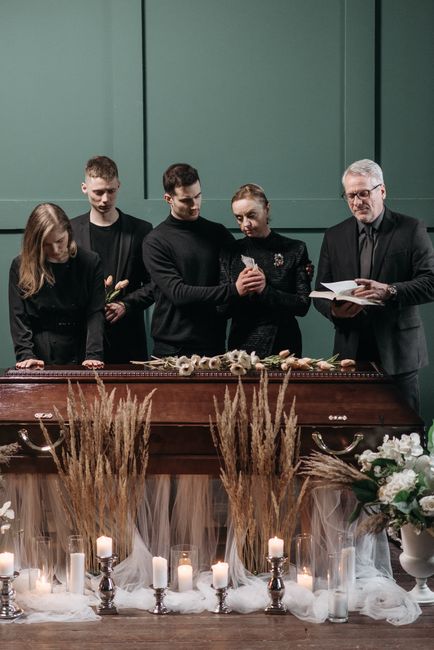 A group of five people gathered around a wooden table displaying a floral arrangement, likely for a condolence or sympathy occasion. The arrangement features a combination of white and cream flowers, 