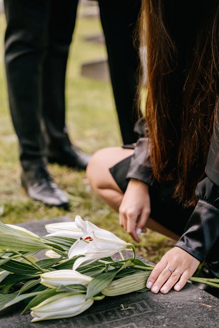A floral arrangement of white lilies with long green leaves is laid on a dark stone surface, likely at a memorial site. The lilies have large, trumpet-shaped petals with hints of red on the stamens, a
