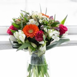 Crafted with an artist's eye by Harold Hill Florist, this bouquet stands as a testament to refined colour harmony and structural grace, echoing the serene landscapes found near Dagnam Park or the vibrant life of local community spaces. Creamy gerberas encircle the arrangement, their petals radiating in soft peach and lively orange around striking brown centers reminiscent of late summer sunlight in Harold Hill. Intermixed are velvety, jewel-toned fuchsia roses, lending luxury and romantic intrigue, while clusters of white alstroemeria and petite spray roses brighten the landscape with their crisp purity. Layered among these, subtle hints of pastel pink and the tiniest white blossoms add nuance and a sense of depth. The bouquet's composition is made complete by robust, glossy green aspidistra leaves, artistically curled to hold the stems in a nurturing embrace-a protective foundation that anchors the effervescent florals above. Wisps of fine, pointed greenery interlace throughout, building gentle contrast and a fresh, woodland-like texture. Housed in a classic clear glass vase, every vibrant stem is visible beneath the bloom, hinting at the arrangement's hand-tied craftsmanship. Soothed by soft daylight akin to the gentle glow of a Harold Hill afternoon, this display feels at once celebratory and sincere-a perfect centerpiece for reunions, brunches, or thoughtful gestures for a new journey. Every element, from fragrant lilies to lush leaves, sings with attentiveness and local care.